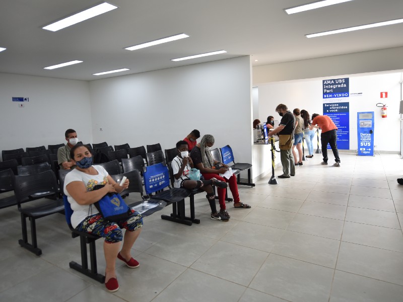 Em primeiro plano, pessoas aguardam sentadas em cadeiras cinzas para serem atendidas. Ao fundo, há uma pequena fila de pacientes recebendo atendimento na recepção da AMA